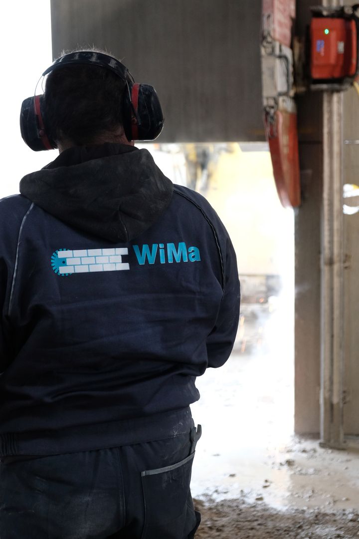 Rückansicht eines Arbeiters in einer blauen Jacke mit 'WiMa GbR' Logo, während er Abrissarbeiten beobachtet.