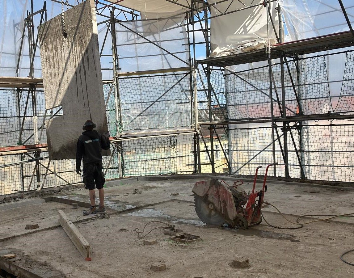 Ein Bauarbeiter steht auf einem Betonboden neben einer großen Kreissäge, umgeben von Gerüsten und Sicherheitsnetzen auf einer Baustelle.