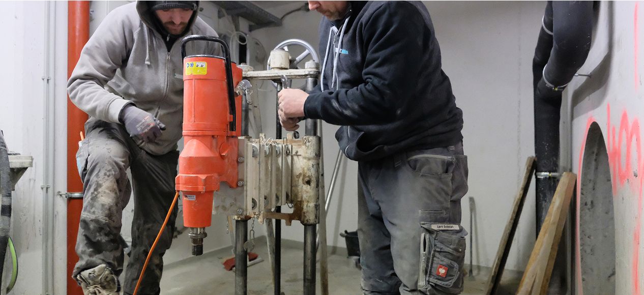 Zwei Arbeiter in grauer Arbeitskleidung montieren oder überprüfen eine Installation mit orangefarbenen Rohren.